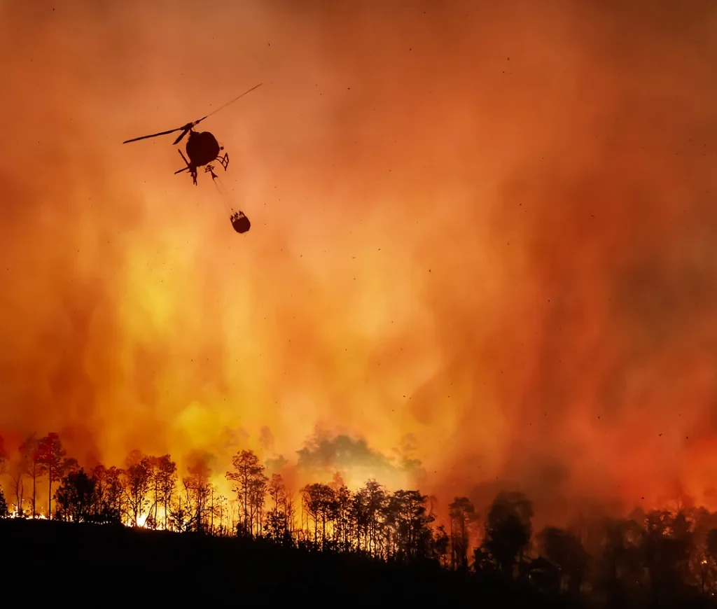 Helicopter with hanging water basket over flame filled sky.