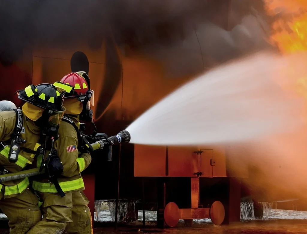 Two firefighters spray water from a fire hose onto flames.