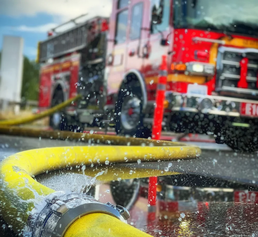 Yellow water hose spraying water from connecting piece with fire truck in background.