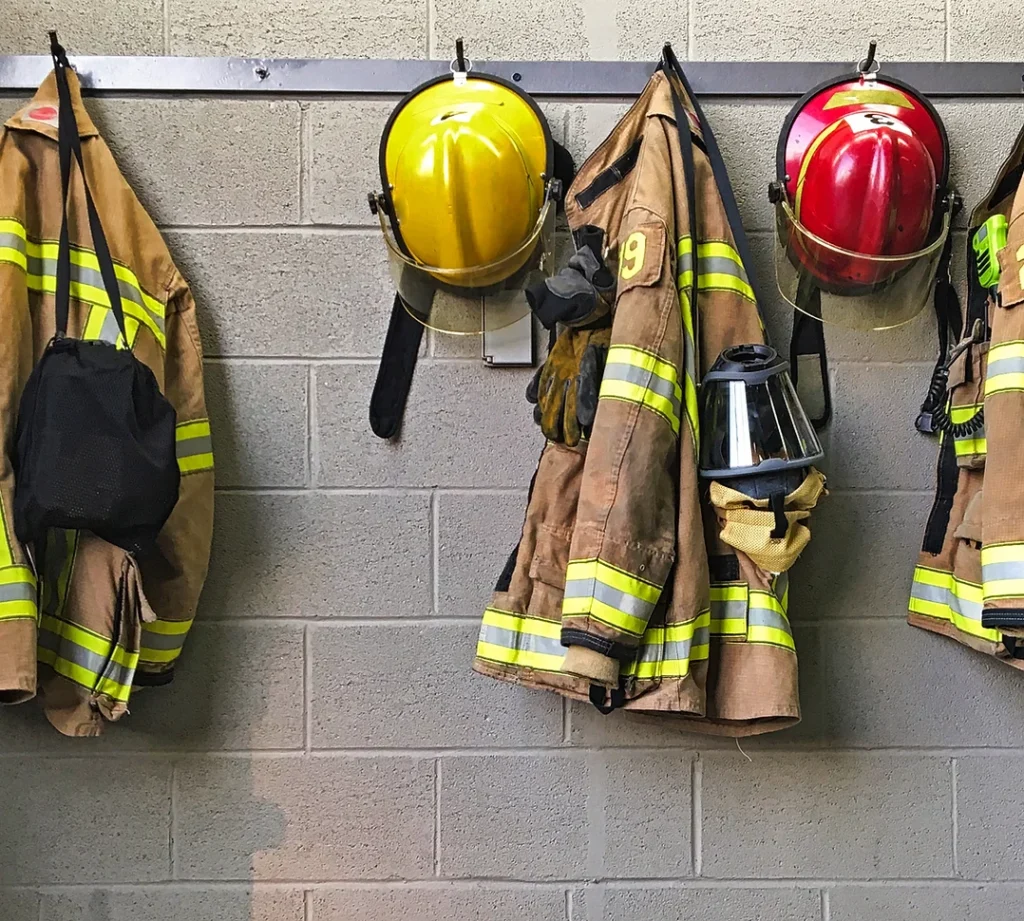 Firefighter coats and helmets hung on hooks on cinderblock wall.