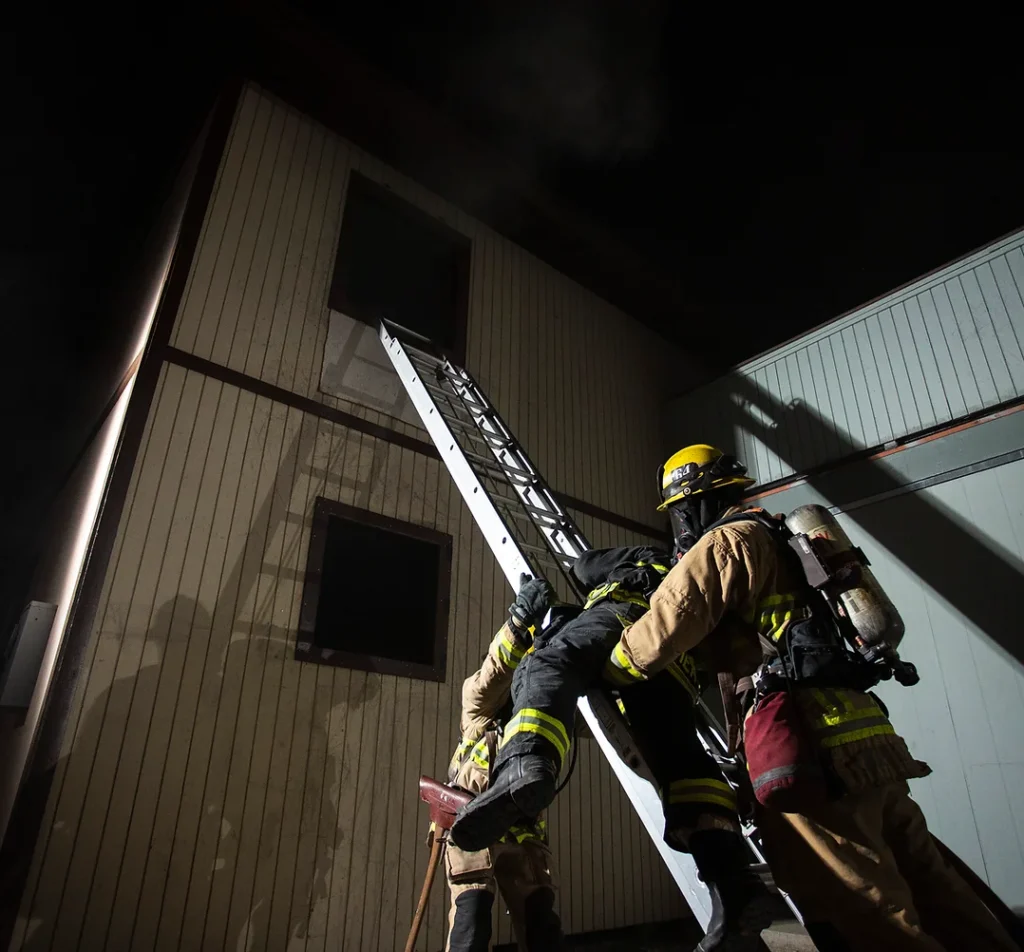 Firefighters climbing on a ladder extended into a building's second floor window.